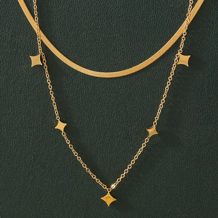 A close-up of a layered gold necklace set against a dark green background, highlighting the curved and star charm designs.