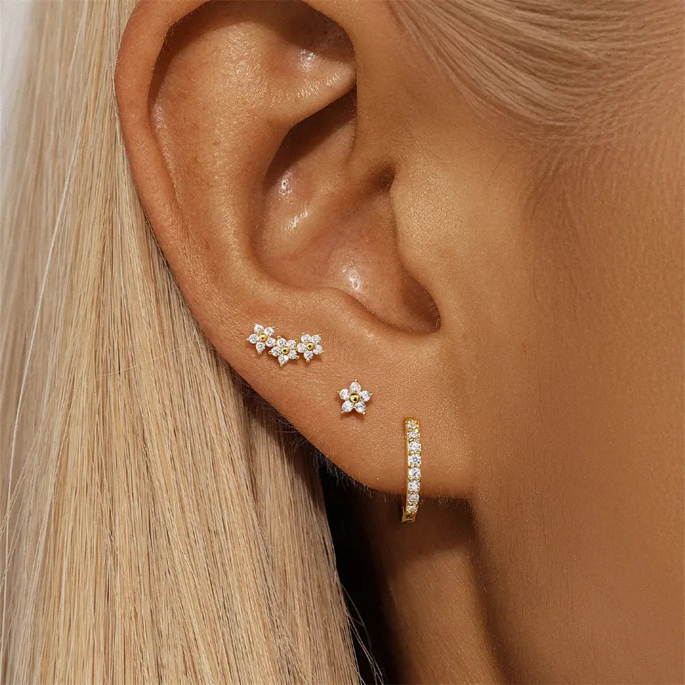 Close-up of a woman's ear wearing three mismatched gold earrings—delicate flower studs and a crystal-adorned huggie hoop.