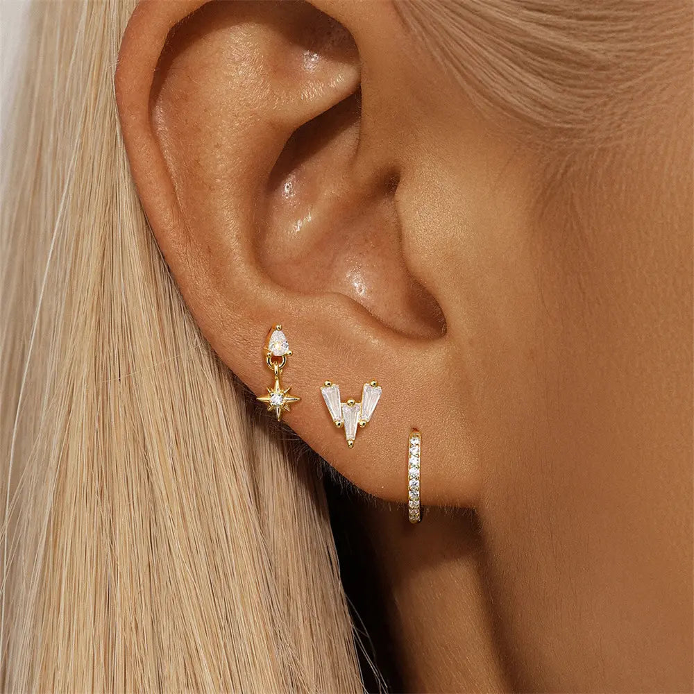 A close-up of a woman's ear showcasing the set of three mismatched gold earrings, including a dangling star earring, a baguette stone trio earring, and a crystal-embellished hoop.