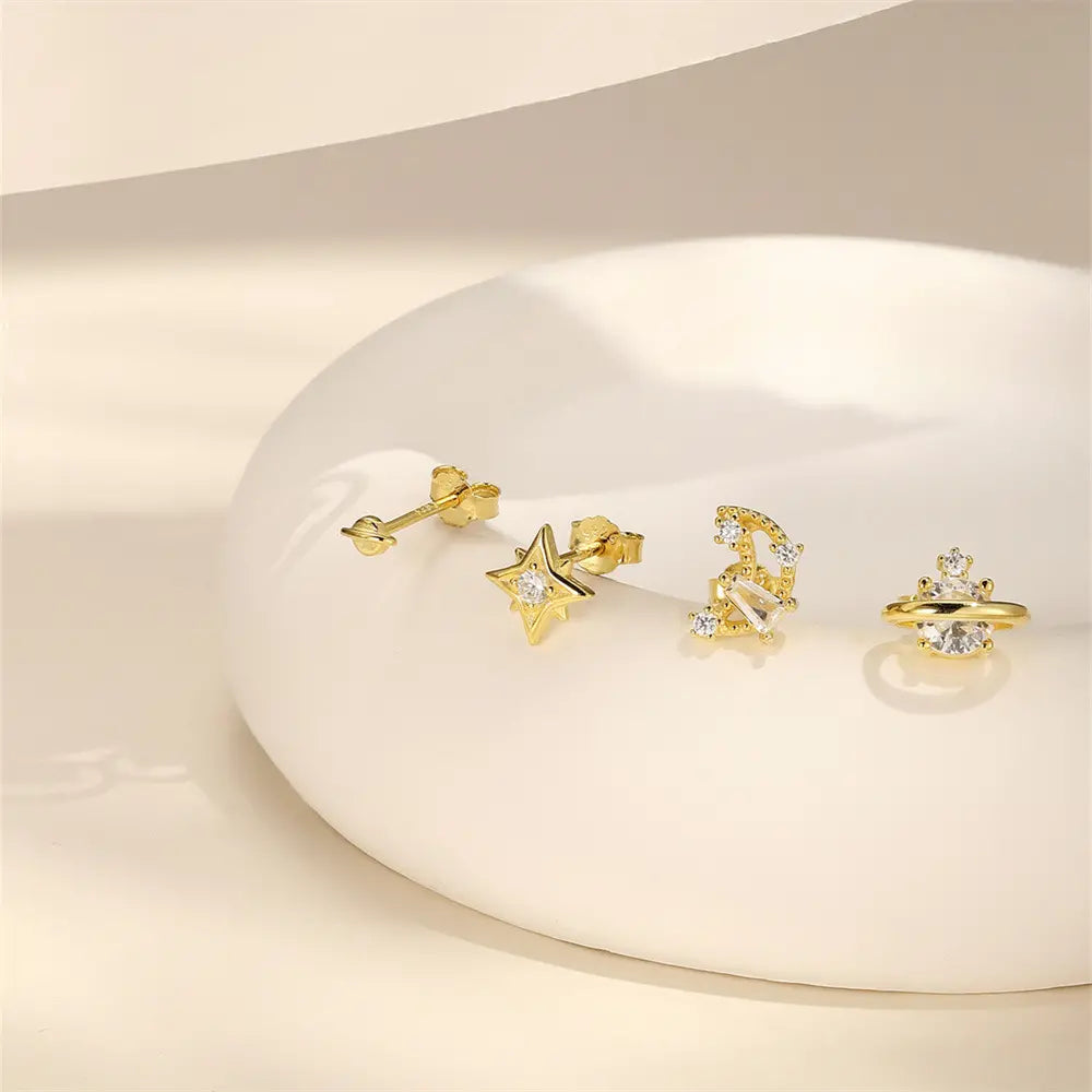 Set of gold celestial-themed stud earrings featuring a star, crescent moon, planet, and minimalist designs with sparkling crystals, displayed on a neutral background.