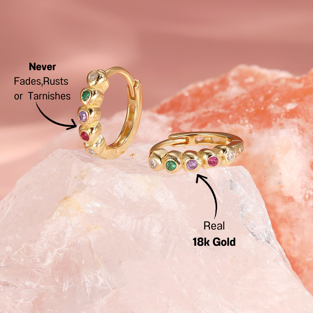 14k-gold-gemstone-huggie-earrings-pink-background.png – Stunning 14k gold huggie earrings adorned with colorful gemstones, displayed on a soft pink background for a chic and feminine vibe.