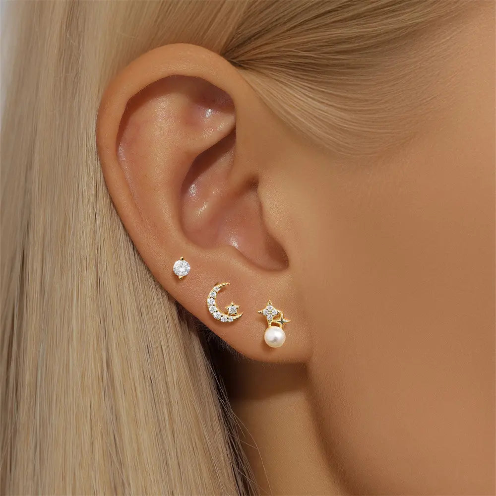 Model wearing gold celestial-themed earrings on the ear, including a round stone stud, a crescent moon stud with crystals, and a gold star with a pearl accent.