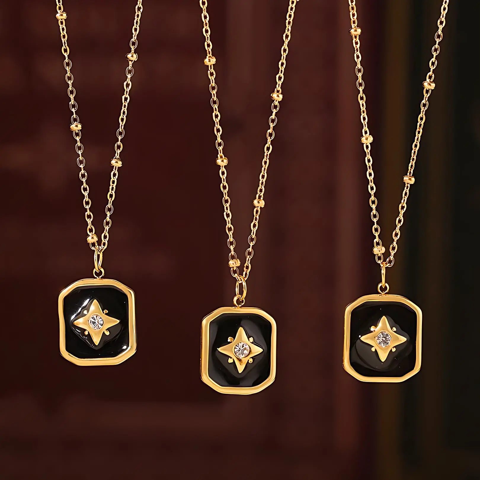 Close-up of three gold necklaces with black enamel rectangular pendants featuring a star and crystal design, displayed against a dark red background.