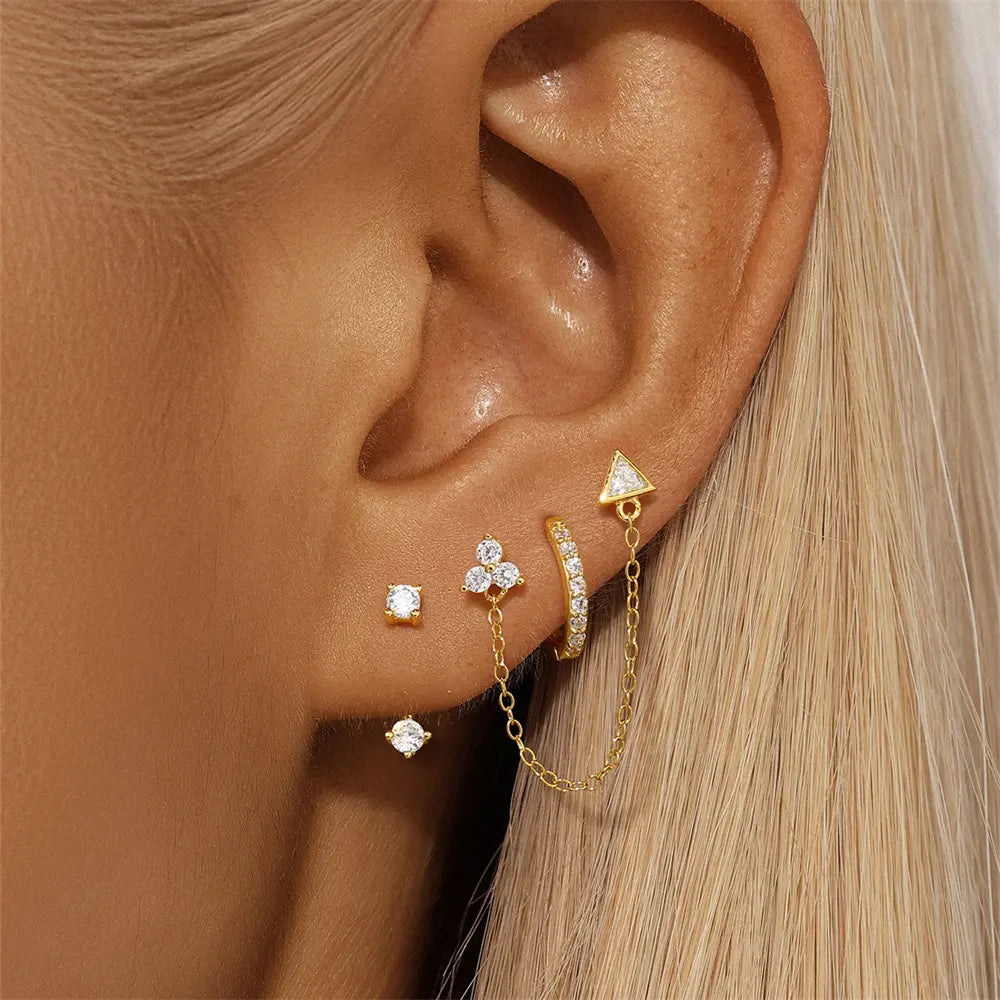 Close-up of an ear wearing mismatched gold earrings, featuring a triangle earring with a dangling chain, a crystal hoop, and studs with unique designs.