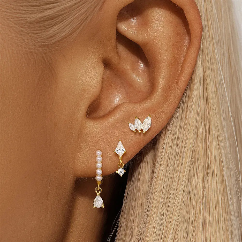 Close-up of a woman's ear adorned with a three-piece gold-plated earring set, including marquise CZ studs, a drop earring, and a pearl huggie hoop with crystal embellishments.