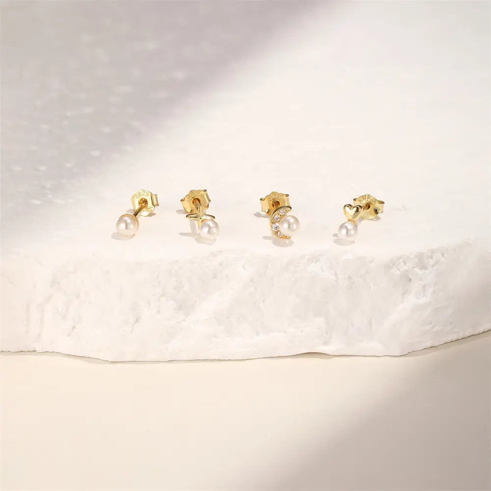 Set of gold stud earrings with pearl accents, featuring moon, star, heart, and round designs, displayed on a textured white background for an elegant presentation.