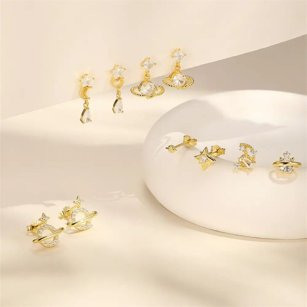 Collection of gold celestial-themed earrings, including planet, moon, and star designs with sparkling crystals, displayed on a soft neutral background.