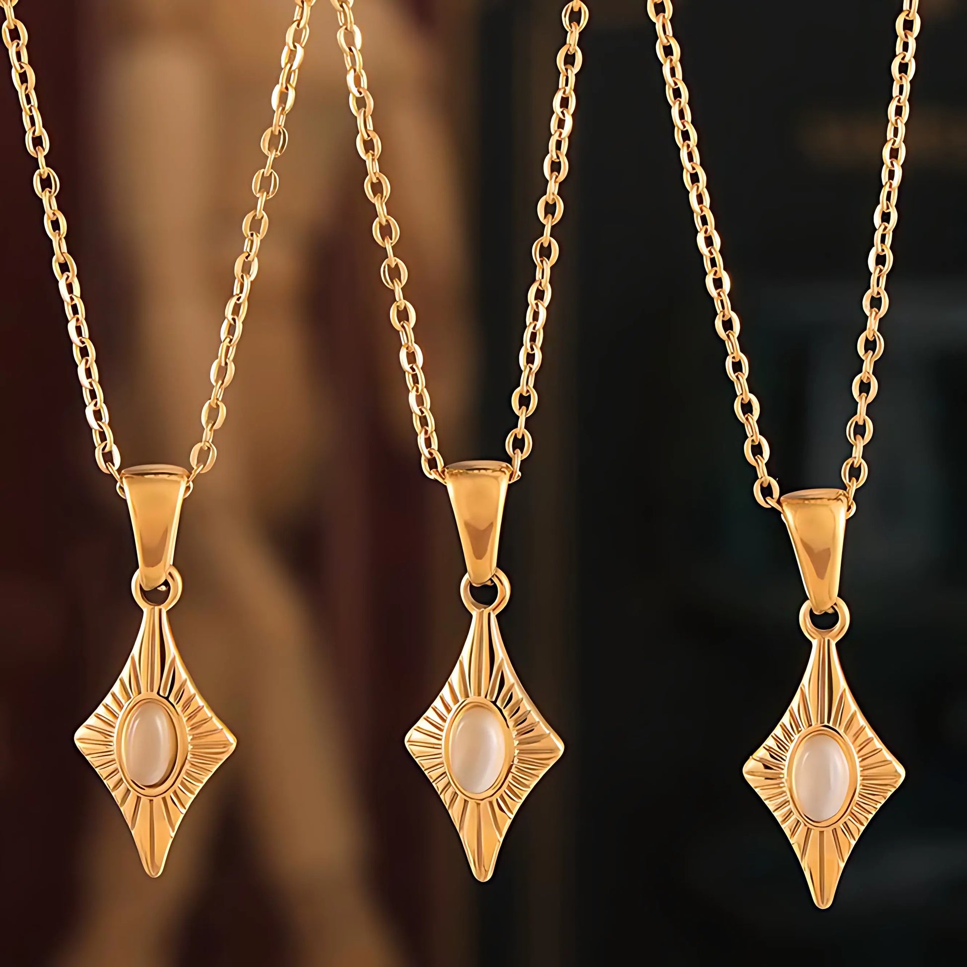 Three diamond-shaped gold necklaces with pearl-like centerstones, suspended on fine gold chains, displayed on a neutral background.