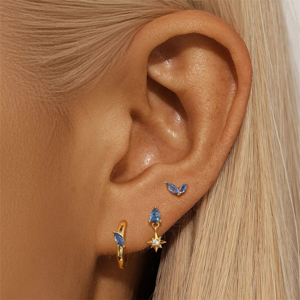 Close-up of a model's ear wearing a gold and blue earring trio, including a drop star stud, marquise stud, and hoop with blue gemstones.