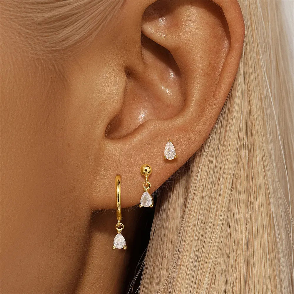 Close-up of a woman's ear wearing three gold earrings: a stud, a dangling charm with a teardrop stone, and a sleek gold hoop with a matching teardrop pendant.