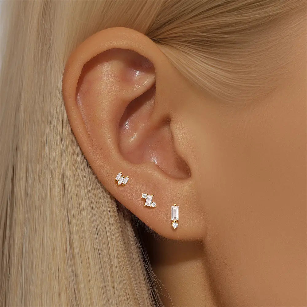 Close-up of a woman's ear wearing a set of gold stud earrings with baguette and round-cut crystals, showcasing a minimalist and elegant layered ear stack.