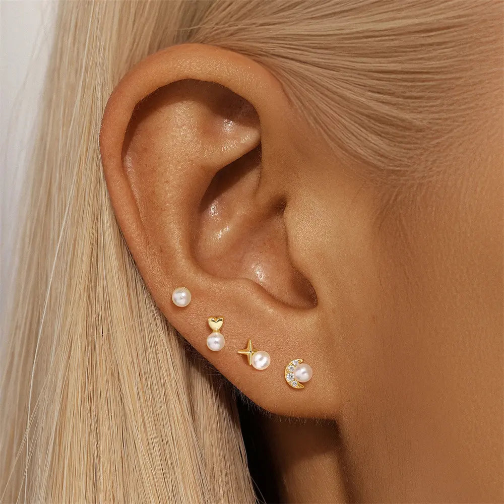 Close-up of a woman's ear wearing a set of gold stud earrings with pearl accents, including moon, star, heart, and round designs, showcasing a chic layered ear stack.